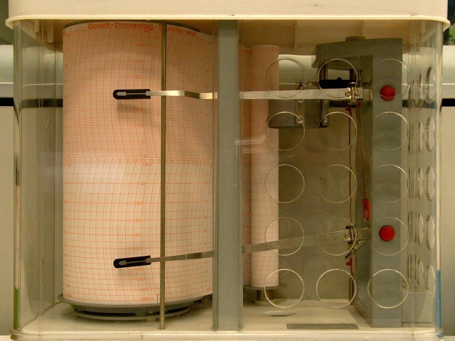 Fotografia przedstawiająca termograf. Jest to plastikowe, przezroczyste naczynie z pokrywką, na której jest metalowa rączka do przenoszenia. W środku naczynia widać: po lewej stronie rolkę papieru stojącą pionowo, po prawej metalowy mechanizm z dwoma wystającymi prętami, które są ruchome, a ich zakończenia dotykają rolki papieru po lewej stronie. 