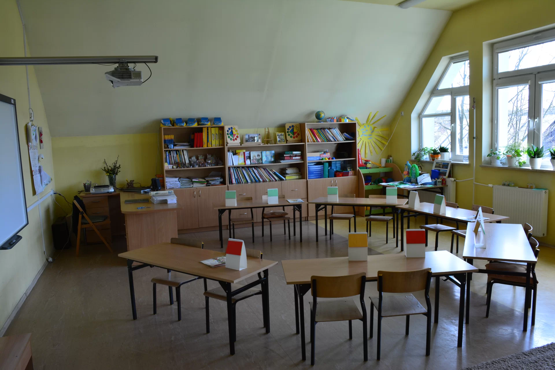 Zdjęcie przedstawia salę klasową. Ławki uczniów poustawiane są w okrąg. Na ścianie zawieszona jest tablica multimedialna, pod sufitem znajduje się rzutnik. Przy biurku nauczyciela stoi rząd półek z książkami i różnymi materiałami dydaktycznymi.