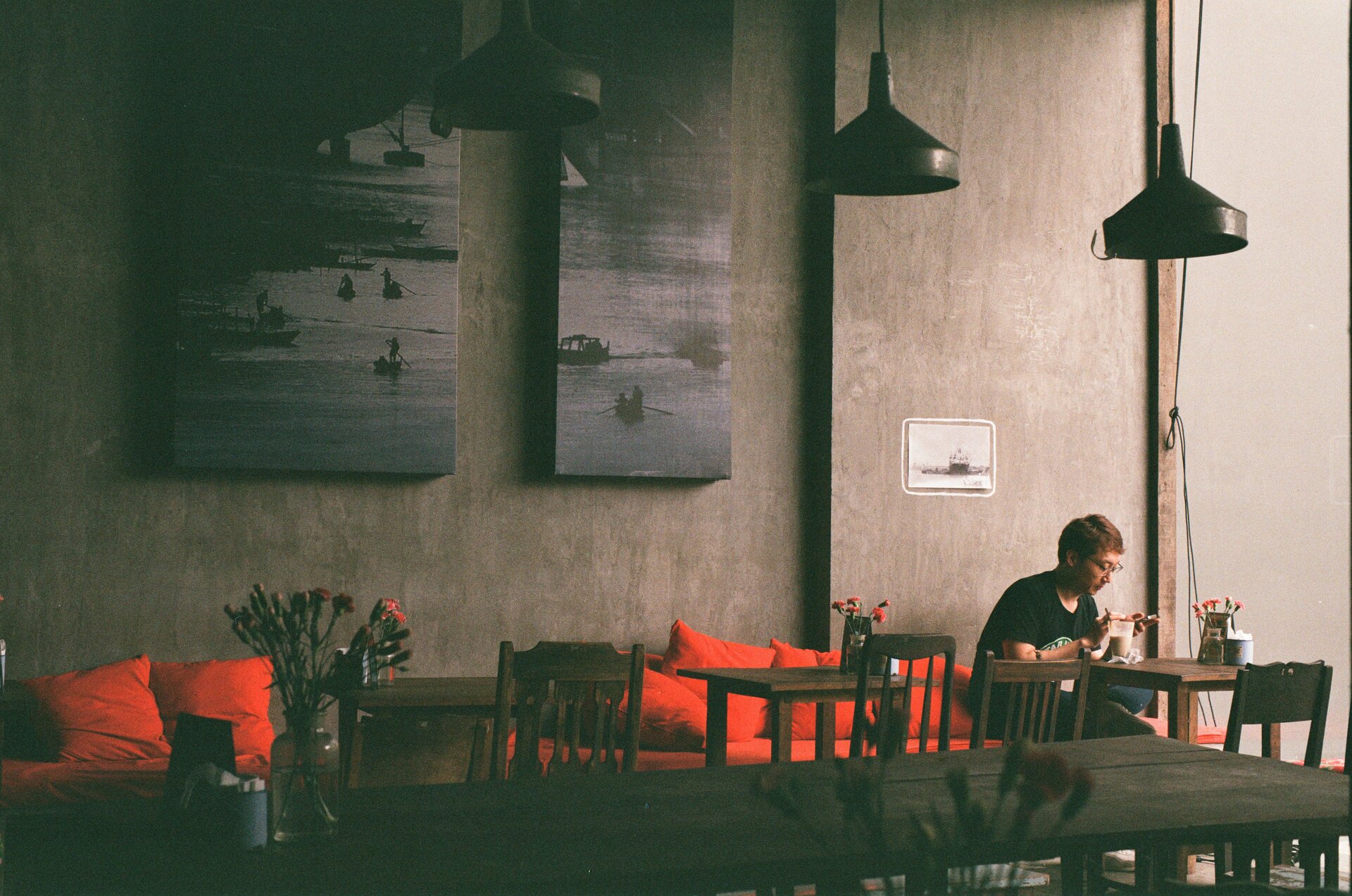 Zdjęcie przedstawia fragment szarej ściany kawiarni. Światło pada z prawej strony, ale okna nie widać. Pod ścianą stoją kanapy z czerwonymi poduszkami, na środku drewniane, proste stoliki z krzesłami. Na stolikach stoją wazoniki z czerwonymi goździkami. Z prawej strony siedzi na kanapie przy stoliku mężczyzna w okularach, sprawdzający coś komórce. Z wysokiego sufitu zwieszają się cztery czarne lampy. Dwa, duże, prostokątne, czarno‑białe obrazy wiszą na ścianie. Mniejszy, biało‑szary, obramowany białą obwódką wisi nad mężczyzną.