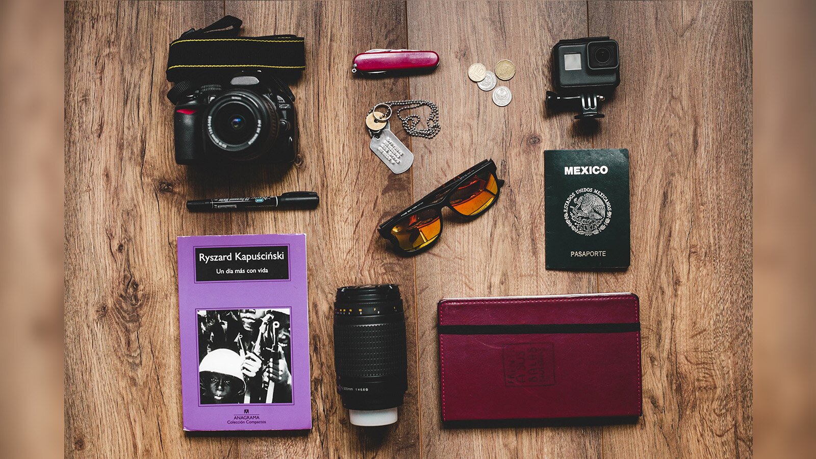 Zdjęcie przedstawia przedmioty potrzebne w podróży. Widoczny jest paszport, aparat fotograficzny, notes, scyzoryk, okulary przeciwsłoneczne, marker i tabliczka znamionowa oraz drobne monety. 