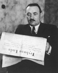 Kliknij żeby powiększyć Mężczyzna z wąsem ubrany jest w garnitur. Siedzi w pomieszczeniu. W rękach trzyma gazetę - Trybunę Ludu. Czyta ją.