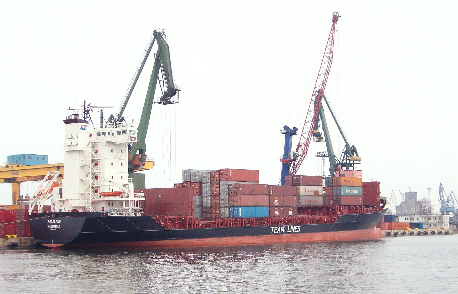 Zdjęcie rozładunku kontenerowca za pomocą żurawi w porcie. W tle inne żurawie i kontenery, na pierwszym planie woda.