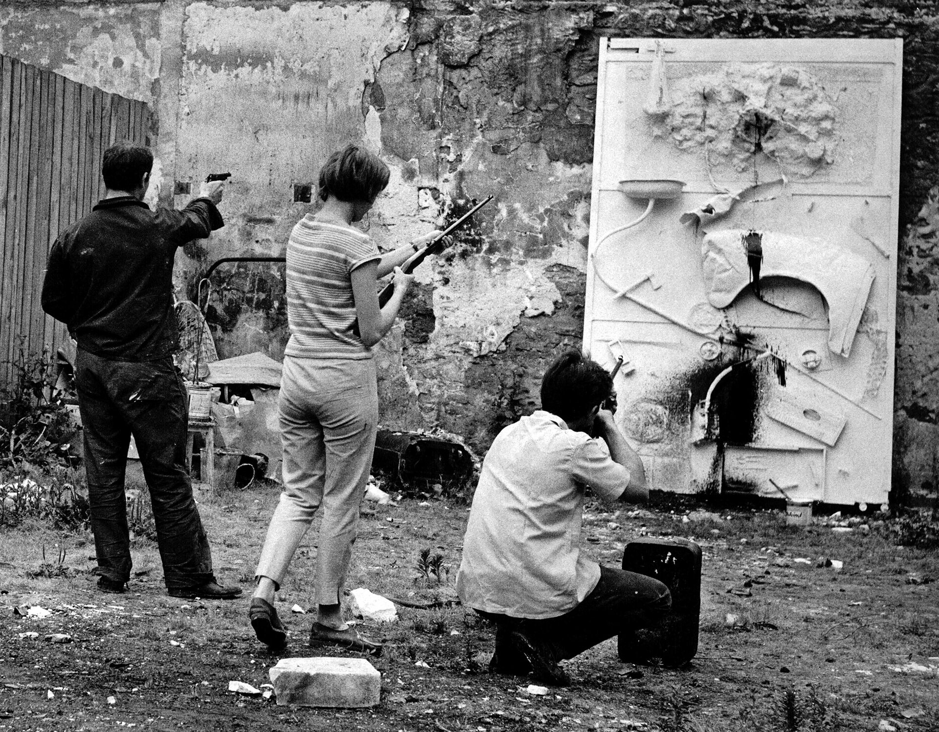 Ilustracja przedstawia fotografię Niki de Saint Phalle i Jean Tinguely podczas strzelania do obrazu-reliefu artystki. Fotografia pochodzi z 1961 roku. Zdjęcie jest czarno-białe.