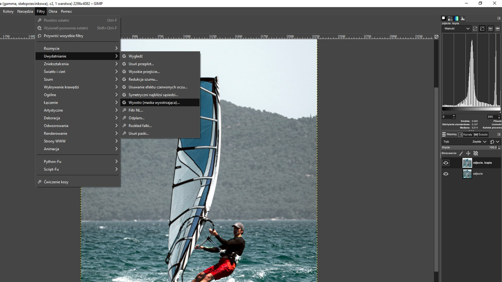 Ilustracja przedstawia okno programu. W obszarze roboczym jest zdjęcie mężczyzny na windsurfingu. Z menu wybrano zakładkę Filtry. Z listy wybrano Uwydatnianie, a następnie Wyostrz (maska wyostrzająca). Po prawej stronie jest panel dotyczący warstw. 
