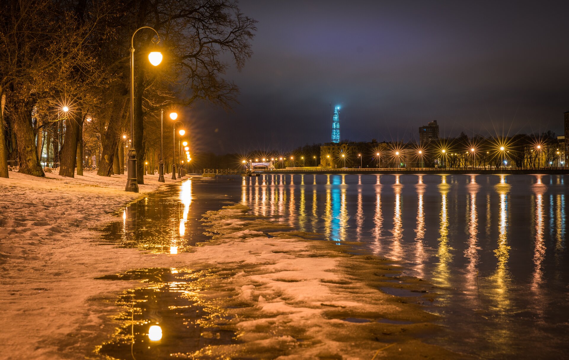 Zdjęcie przedstawia miasto po zmroku. Na alejce widać wodę, obok płynie rzeka.