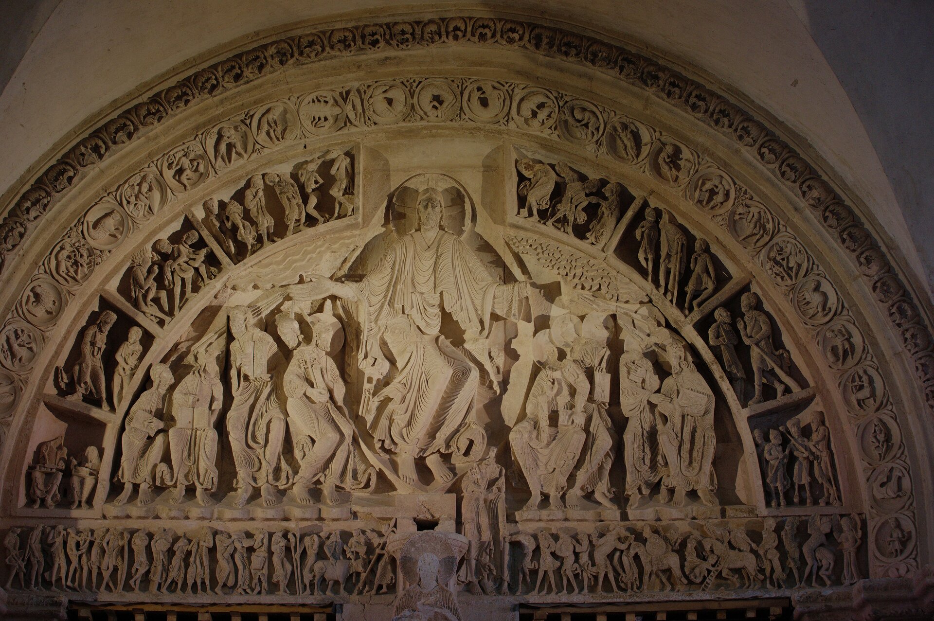 Zdjęcie przedstawia zdobiony rzeźbami tympanon w kształcie półokręgu, wykonany z jasnego kamienia. W centralnej części znajduje się postać Jezusa, z dłońmi rozłożonymi w obie strony. Ma długie włosy i zarost. Wokół jego głowy umieszczono aureolę z krzyżem. Ma na sobie długie szaty; wyrzeźbiono na nich cienkim dłutem linie układające się w fale i okręgi. Po obu stronach Jezusa znajdują się postacie. Niektóre również mają aureole. Wszystkie postacie siedzą, mają głowy zwrócone w różne strony, niektóre mają książki lub inne przedmioty w dłoniach. Scena jest otoczona kolejnym półokręgiem, gdzie zostało wydzielone liniami osiem różnych scen. Na każdej znajduje się przynajmniej dwie postacie. Następny okalający półokrąg przedstawia wiele stworzeń, takich jak: pół człowiek i pół koń z łukiem, stworzenie przypominające raka, pół ludzie z rybimi ogonami i inne. Ostatni półokrąg zdobiony jest florystycznym wzorem. Na dole, w linii poziomej są ustawione blisko obok siebie postacie z koszami, tarczą, długimi narzędziami oraz krowa czy konie.
