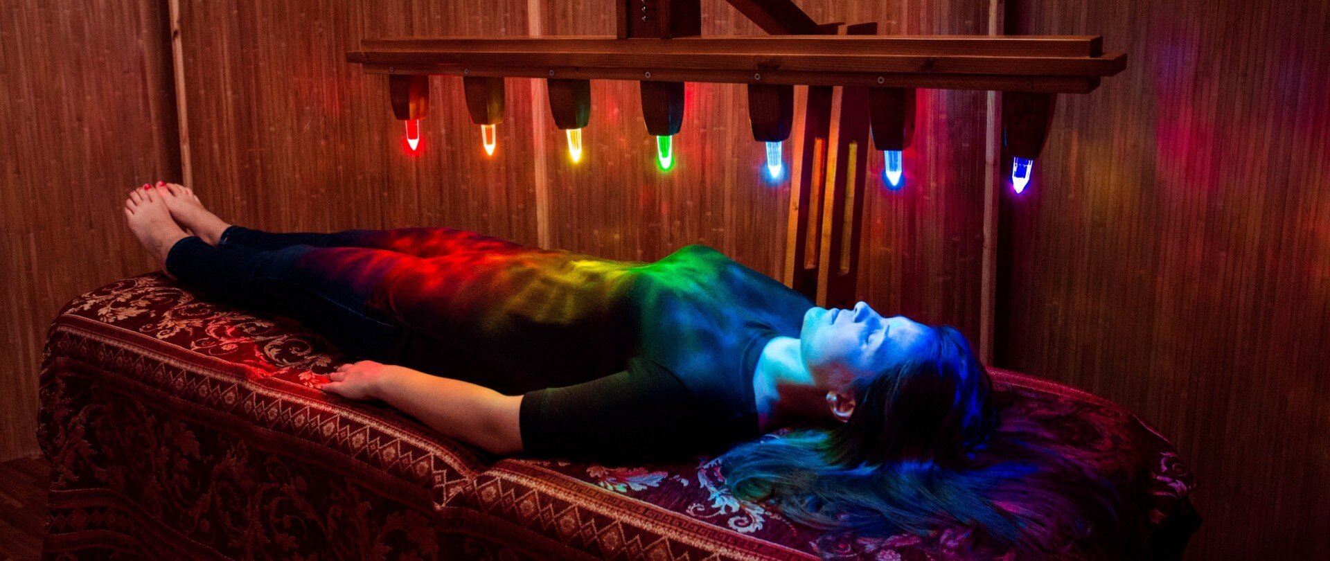 Fotografia przedstawia kobietę o ciemnych włosach, w czarnym stroju, która leży na łóżku wyłożonym wzorzystą, ciemną narzutą. Kobieta leży na plecach, a ręce trzyma wzdłuż ciała. Znajduje się w pomieszczeniu o drewnianych ścianach. Nad nią zwieszony jest prostokątny, drewniany uchwyt, z którego wystają różnokolorowe żarówki. Od lewej strony fotografii jest to czerwona, pomarańczowa, żółta, zielona, jasnoniebieska, ciemnoniebieska oraz fioletowa żarówka.
