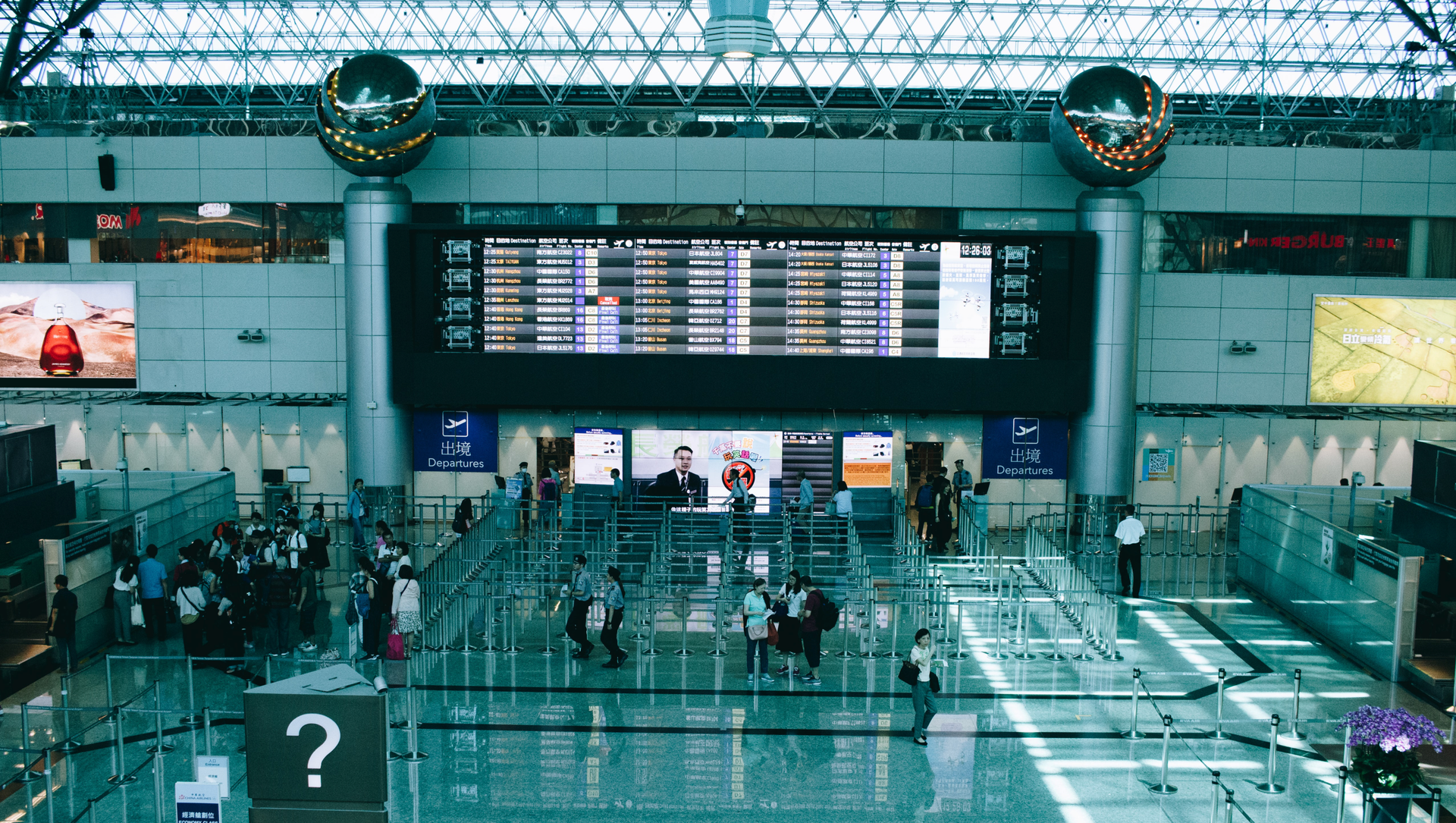 Zdjęcie przedstawia w szerokim kadrze halę lotniska. Widoczni są ludzie oczekujący w kolejce do kontroli. Widoczna jest duża tablica przylotów i odlotów.