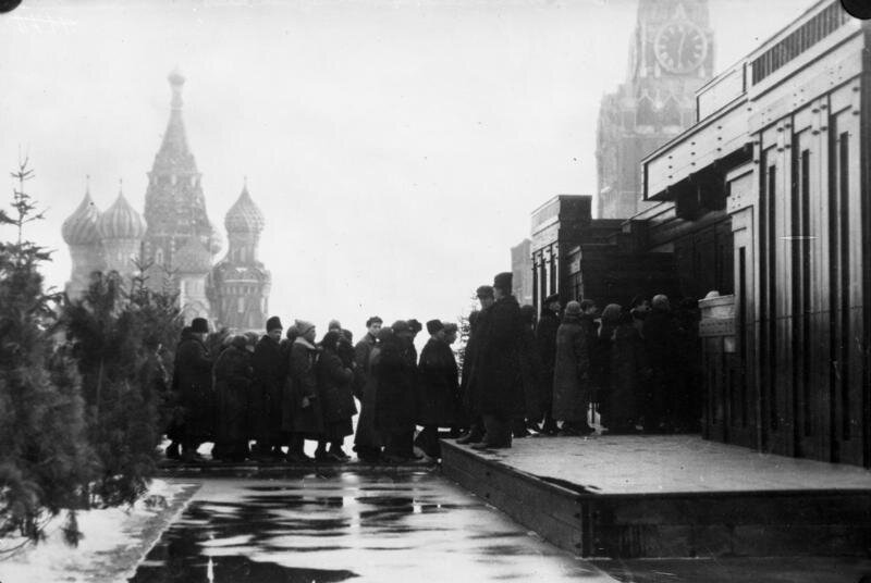 Zdjęcie przedstawia Plac Czerwony i wejście do Mauzoleum Lenina w Moskwie. Prezentuje ludzi stojących w kolejce do Mauzoleum. W tle widać cerkiew.