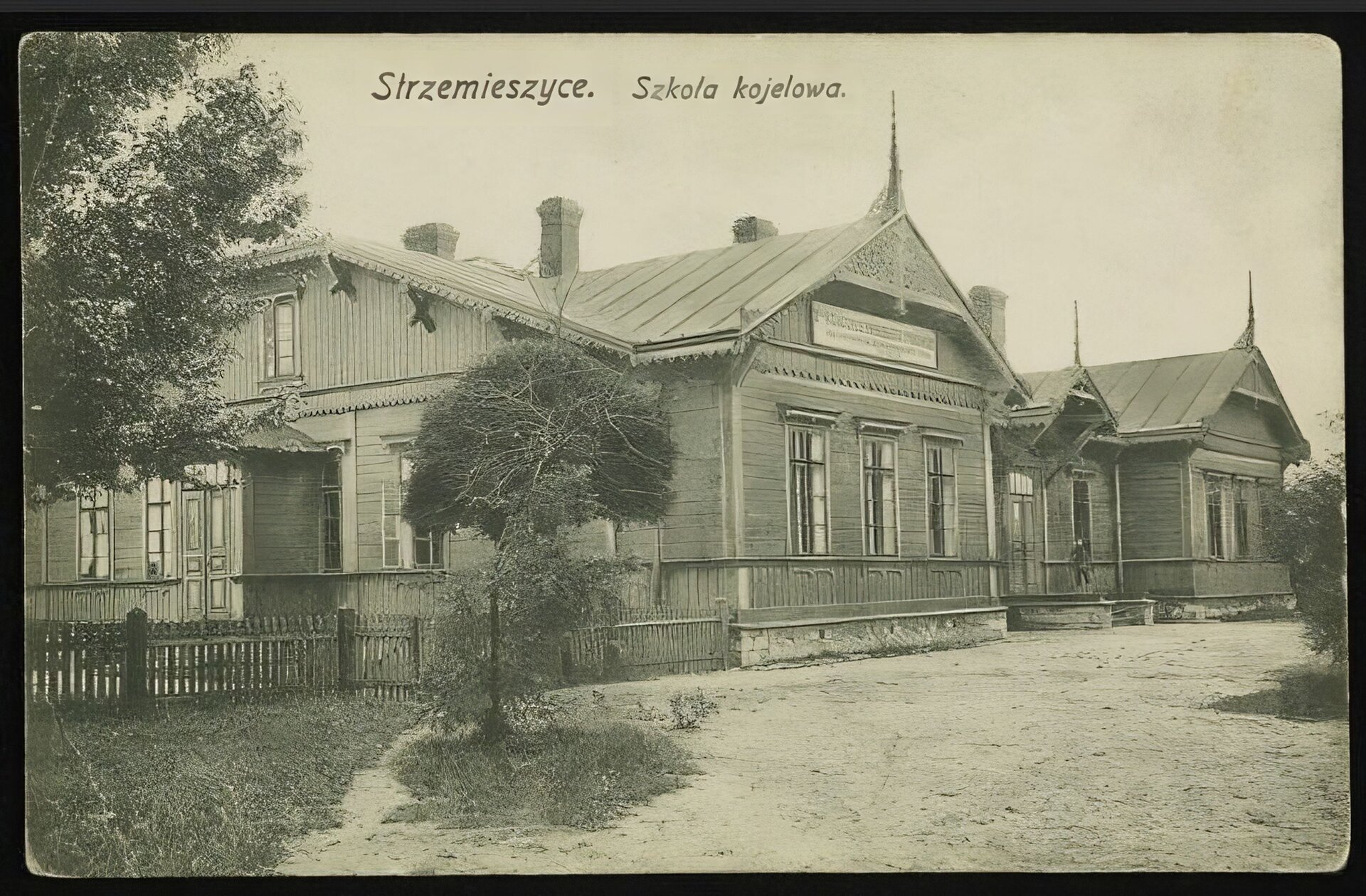Kliknij, aby powiększyć. Zdjęcie przedstawia fragment jednopiętrowego, drewnianego budynku. Składa się z kilku połączonych ze sobą części. Każda z nich posiada okna i spadzisty dach zakończony ozdobnymi rzeźbionymi, spiczastymi elementami . Z lewej strony widać ganek i wejście do budynku. Wokół budynku stoi płot i kilka drzew. Przed płotem biegnie droga. Na zdjęciu u góry jest napis: Strzemieszyce. Szkoła kolejowa.