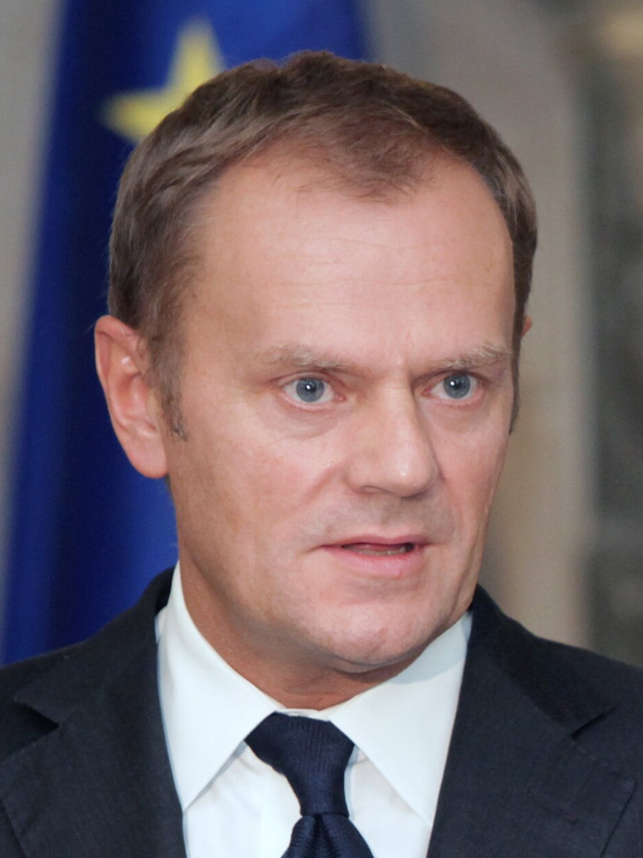 Fotografia przedstawiająca dojrzałego mężczyznę. Postać przedstawiona jest od piersi w górę. Ukazuje prawy półprofil. Polityk ma owalną twarz, gładko zaczesane włosy, zakola, przerzedzone brwi, oczy lekko zasłonięte powiekami, prosty nos, wąskie, lekko otwarte usta. Ubrany jest w garnitur, ma zawiązany pod szyją krawat. W tle flaga Unii Europejskiej.