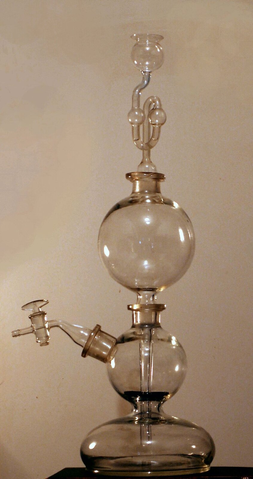 Ilustracja przedstawiająca aparat Kippa zbudowany z trzech okrągłych komór szklanych umieszczonych jedna nad drugą i połączonych za pomocą szklanych szlifów zapewniających szczelność aparatury. Górna komora łączy się z za pomocą szklanej rurki z dolną komorą, która to ma dostęp do komory środkowej dzięki oddzielającej komory płycie retencyjnej. W środkowej komorze z reguły umieszcza się ciało stałe. Środkowa komora posiada kranik w górnej części umożliwiający odpływ gazu. Poprzez górną komorę doprowadza się ciekły reagent, na przykład kwas.