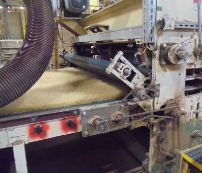 Zdjęcie przedstawia igłowarkę - maszynę do produkcji wyrobów z włókna kokosowego. Stoi w hali zakładu produkcyjnego. Po lewej stronie taśma z włóknem kokosowym, które wprowadzane jest pomiędzy wałki. Nad taśmą z włóknem elastyczna, karbowana rura. 