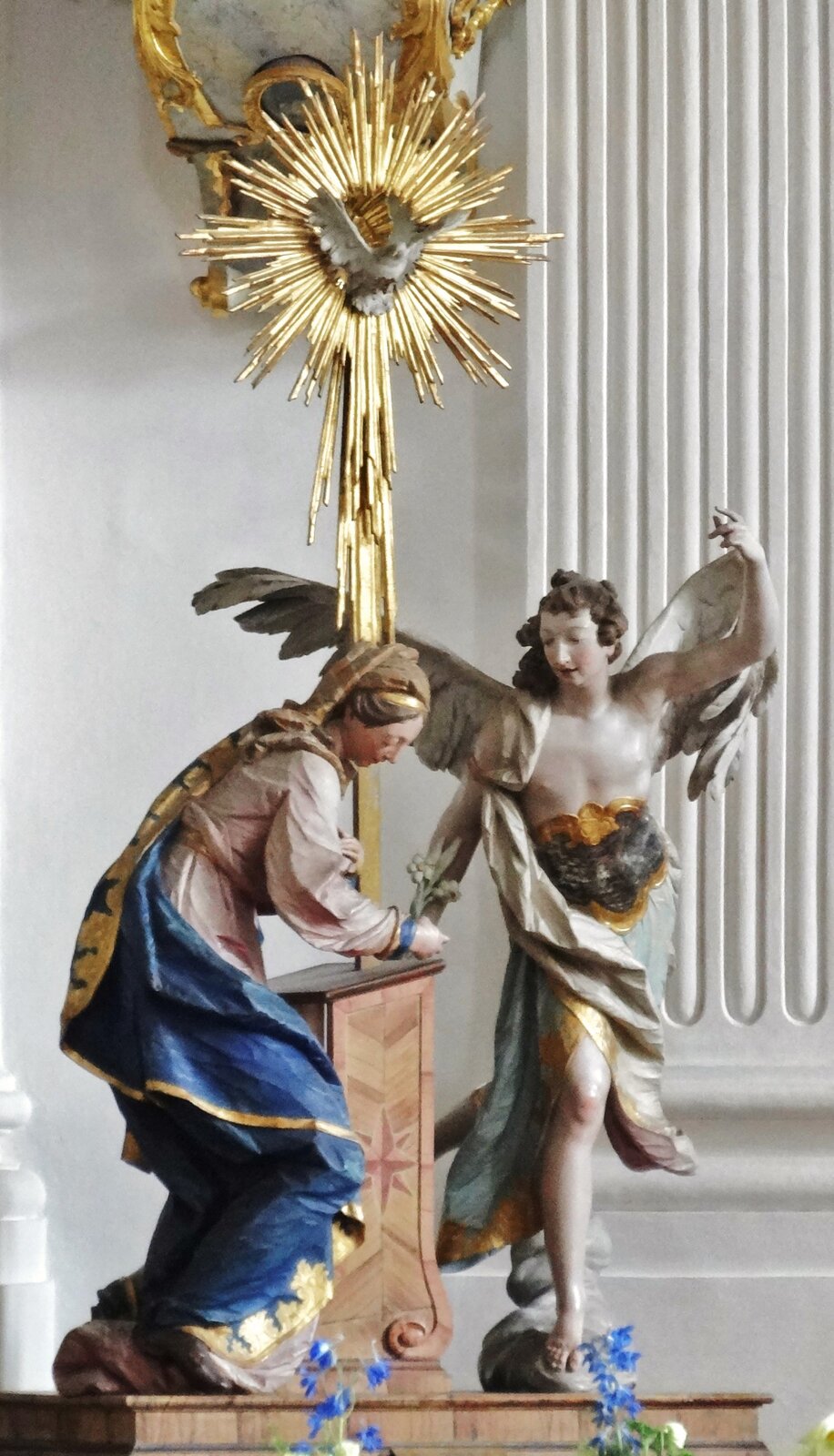 Ilustracja przedstawia rzeźbę „Zwiastowanie z kościoła w Weyarn”. Rzeźba przedstawia postacie obok monstrancji. Po prawej stronie stoi postać Anioła o długich skrzydłach. Z lewej strony widoczna jest kobieta trzymająca w prawej dłoni kwiaty. Po środku znajduje się monstrancja koloru złotego.