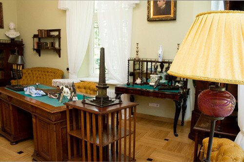 Fotografia przedstawia wnętrze pomieszczenia. Przy ścianie po lewej stronie zdjęcia stoi drewniany regał. Stoi na nim rzeźbione popiersie mężczyzny. Obok stoi kanapa. Za kanapą umieszczone jest okno. Pośrodku pomieszczenia stoi ciężkie, drewniane biurko. Na blacie biurka leżą artykuły piśmiennicze. Stoi na nim lampa i ramki ze zdjęciami. Obok biurka stoi dość wysoka, drewniana kolumna. Na blacie stoi rzeźba przedstawiająca obelisk. Na ścianie wisi portret młodego mężczyzny. Po prawej stronie stoi wysoka lampka.