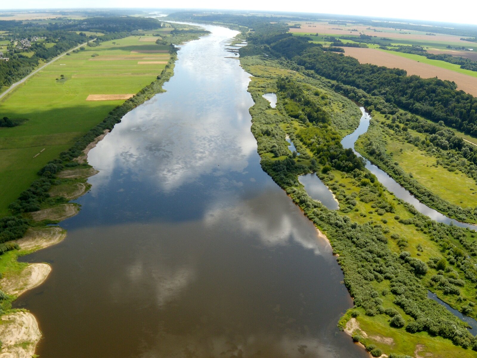 Rzeka Niemen, fotografia lotnicza. Pośrodku szeroka rzeka. Wokół zielona trawa i drzewa, płaski teren.