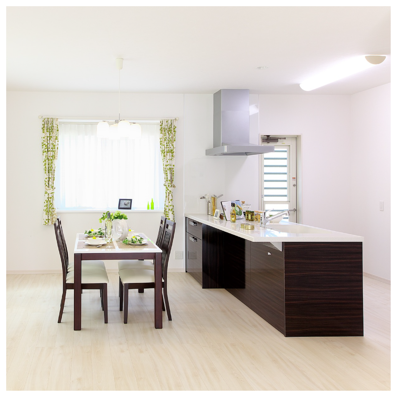 Widoczna jest kuchnia z jadalnią. W centrum po oknami stoi stół z czterema krzesłami. Po prawej stronie urządzenia kuchenne.