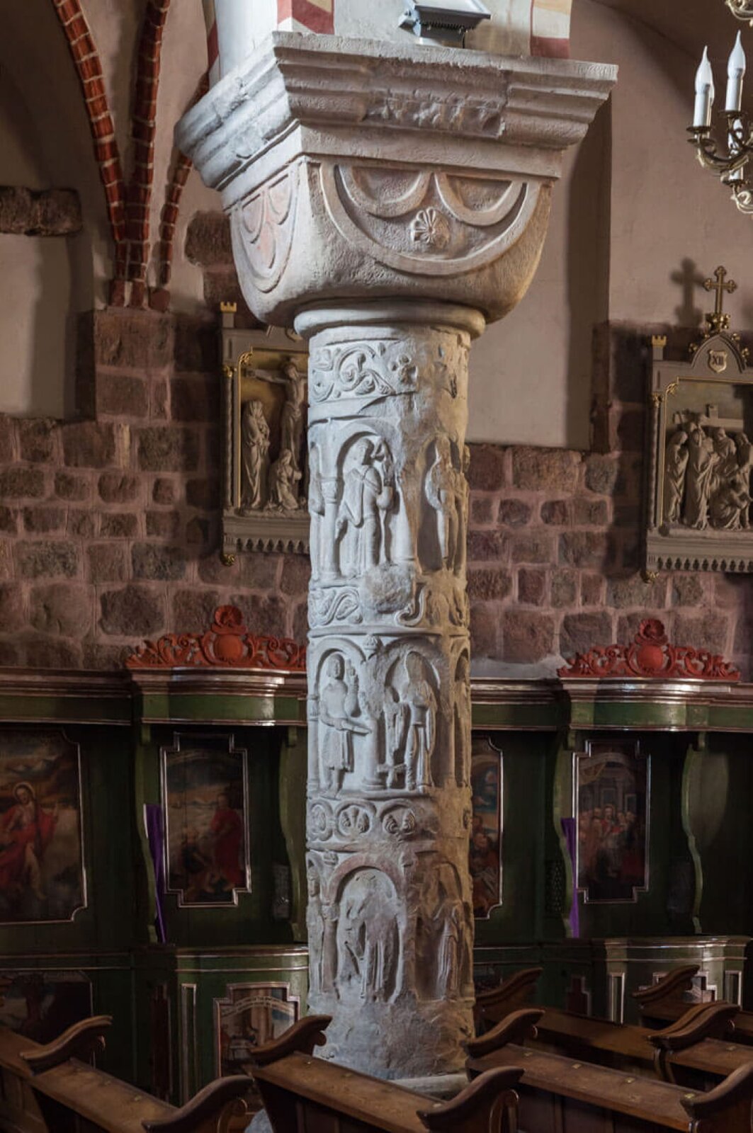 Fotografia przedstawia wnętrze budynku, którego konstrukcja została oparta na kolumnach z głowicami kostkowymi z półkolami. Kolumny wraz z głowicami zostały wykonane z szarego kamienia. Kształt głowic przypomina graniastosłup prosty o podstawie prostokąta i zaokrąglonych ścianach. Głowice posiadają zdobienia - na ich ścianach wymodelowano trzy półkola - jedno duże, w którym umieszczono dwa mniejsze. Pod mniejszymi półkolami ornament przypominający kwiat. Kolumna posiada zdobienia - płaskorzeźby, które przedstawiają postacie ludzkie - część z nich stoi z bronią. Wnętrze budynku ozdobione jest obrazami z wizerunkiem świętych.
