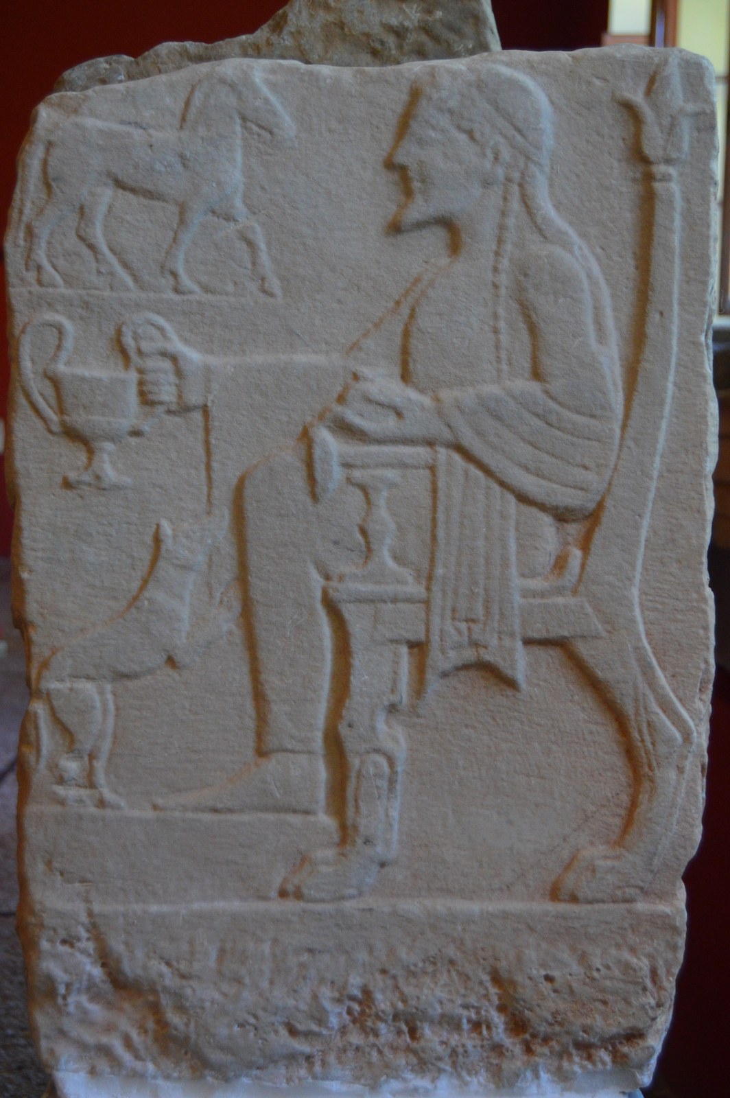 Ilustracja przedstawia fragment płaskorzeźby przedstawiającej mężczyznę siedzącego na krześle, trzymającego w ręce puchar. Ponad wyciągniętą ręką mężczyzny znajduje się płaskorzeźba konia, poniżej znajduje się płaskorzeźba kota.