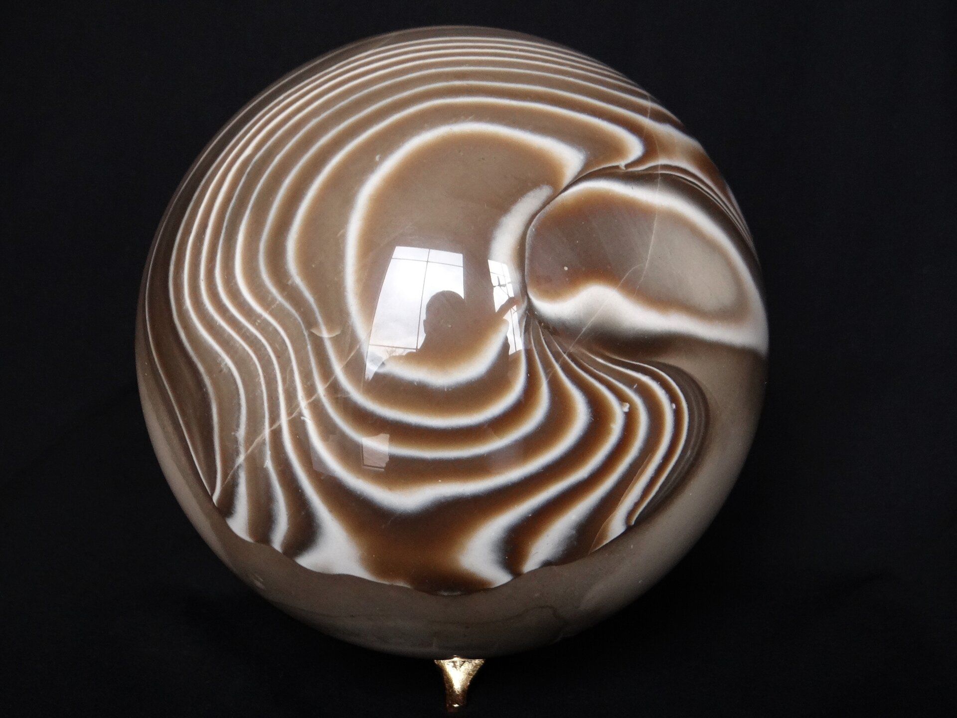 Zdjęcie przedstawia prawie idealną kulkę z krzemienia pasiastego o bardzo nieregularnym, brązowo białym wzorze zanikającym w dolnej części. Kulka jest wygładzona na wysoki połysk i ma widoczne rysy. W samym środku od jej powierzchni odbija się kształt okna i sylwetka osoby wykonującej zdjęcie.