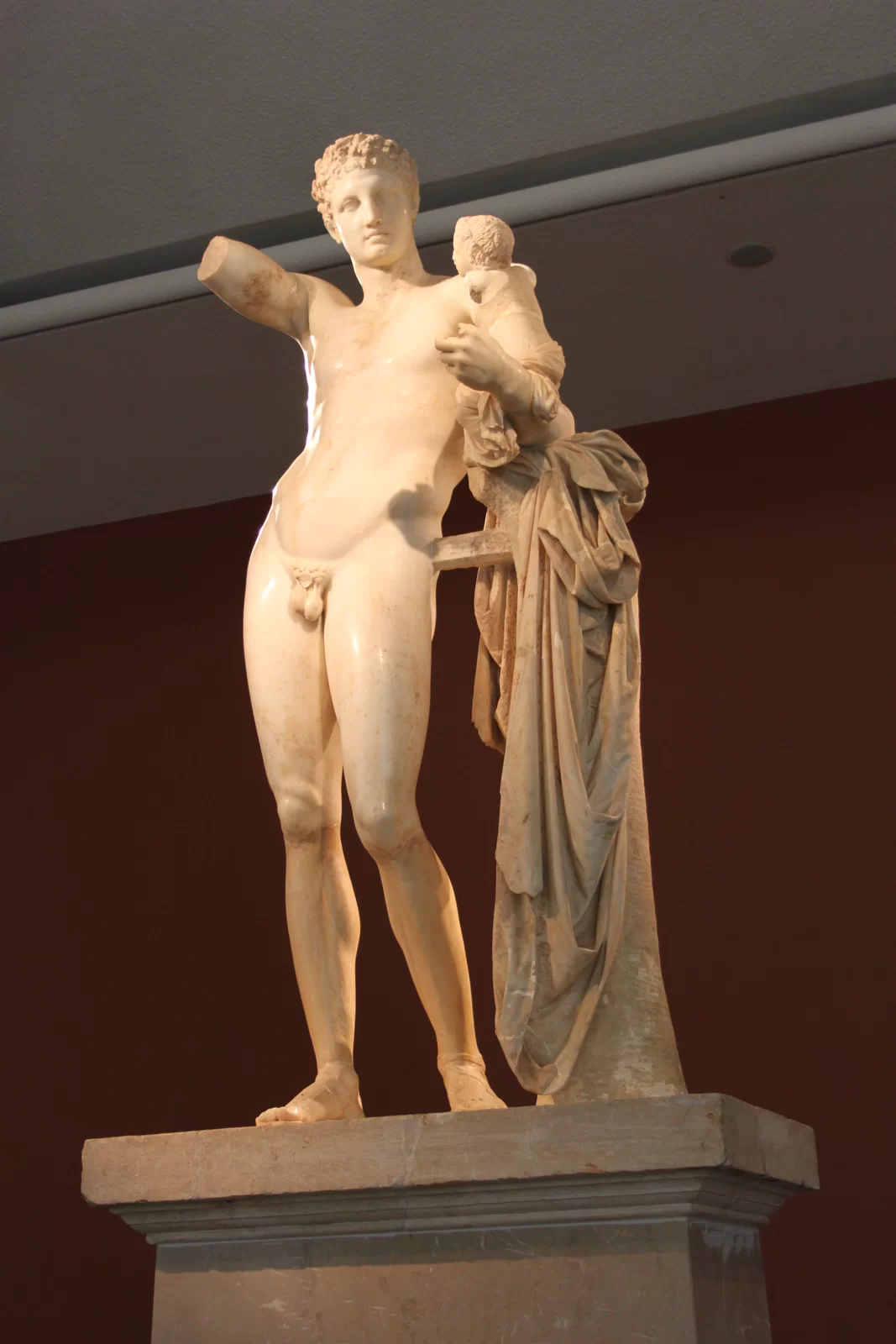 Zdjęcie przedstawia rzeźbę pod tytułem „Hermes i mały Dionizos”. Rzeźba jest wykonana z jasnego, błyszczącego kamienia. Składa się z wizerunku nagiego, stojącego mężczyzny, który na swym lewym ramieniu trzyma małe dziecko, a pod dzieckiem znajduje się długa, obszerna, opadająca szata. Ciało mężcyzny jest umięśnione. Stoi na prawej, wyprostowanej nodze, a opiera się palcami na lewej. Na łydkach rysują się mięśnie. Tors ma lekko odchylony do tyłu, ukazując mięśnie na brzuchu. Mężczyzna patrzy w lewą stronę. Ma krótkie, kręcone włosy, małe usta, szczupły nos, nieodstające uszy. Dziecko, które trzyma na zgiętym lewym ramieniu, siedzi wyprostowane. Ma krótkie, kręcone włosy. W pasie ma zawiązaną szatę, która opada w dół, aż do samej ziemi. Rzeźba jest wybrakowana w niektórych miejscach - mężczyzna nie ma prawej ręki od łokcia, brakuje mu palców u lewej dłoni, a dziecko nie ma rąk. Jest ustawiona na kamiennej kolumnie.
