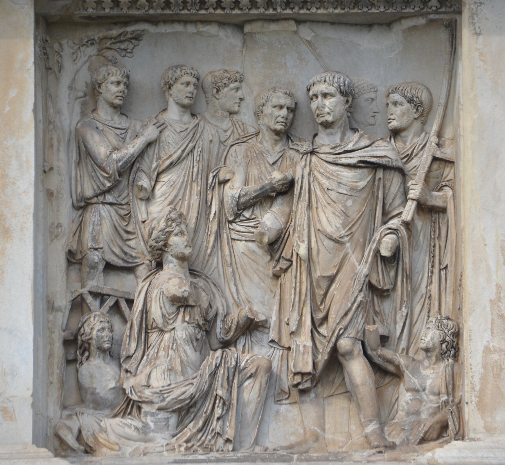 Na ilustracji przedstawiono fragment kolumny Trajana z wyrzeźbionymi w jasnym kamieniu postaciami. Wszyscy mają na sobie długie, zwiewne szaty. W centrum uwagi, umieszczony w prawej części rzeźby został mężczyzna zwrócony w przód, patrzący przed siebie. Obok niego, po lewej stronie, klęczy inna postać. Wokół stoi jeszcze sześć innych mężczyzn - im dalej zostali przedstawieni, tym mniej wystają poza powierzchnię rzeźby, są niemal płascy. Po bokach, na dole siedzą jeszcze dwie postacie. Ręce postaci w większości nie zachowały się, odpadły od łokcia w dół.