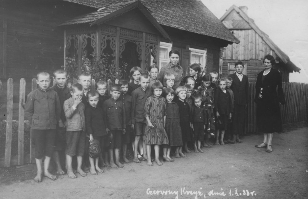 Fotografia przedstawia grupę dzieci z nauczycielką stojącymi przed małym budynkiem ogrodzonym płotkiem. Grupa jest nieliczna. Dzieci są ubrane w zwykłe ubrania i nie mają butów.