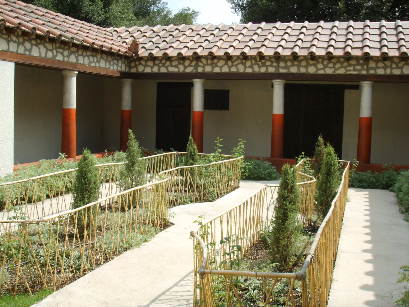 Fotografia przedstawia ogród na wewnętrznym dziedzińcu rzymskiego domu. Rośliny oddzielone są od betonowych chodników barierkami. Konstrukcja domu wsparta jest na kolumnach, które podtrzymują dach budynku.