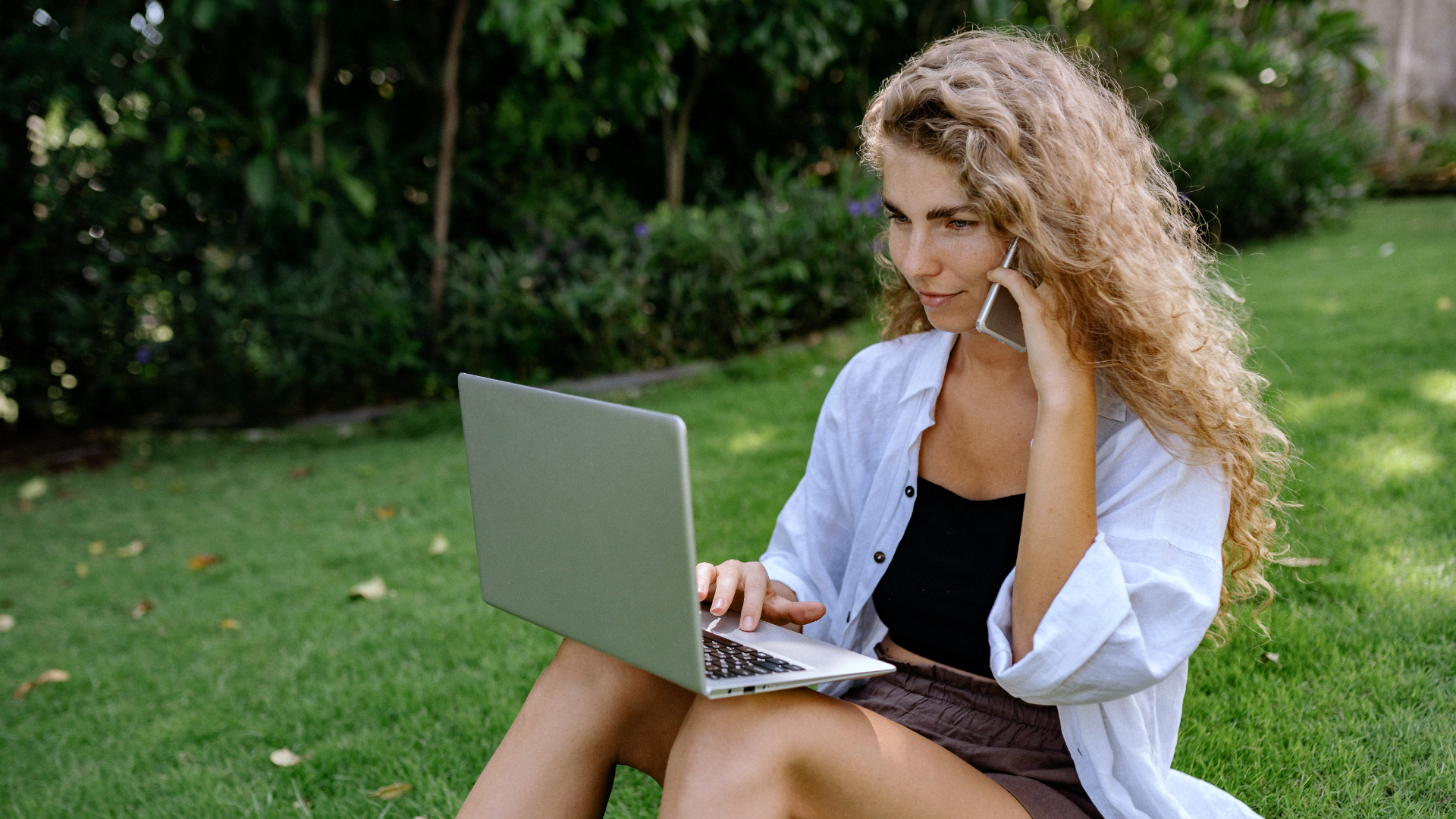Zdjęcie przedstawia młodą kobietę z długimi, kręconymi włosami. Kobieta siedzi na trawie, na kolanach trzyma otwartego laptopa. Lewą ręką przy uchu trzyma telefon komórkowy.