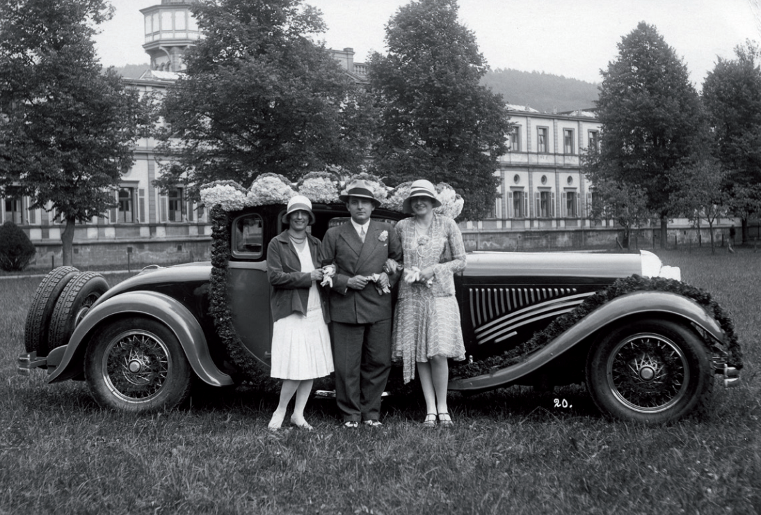 Ilustracja przedstawia mężczyznę stojącego w otoczeniu dwóch kobiet na tle samochodu z lat trzydziestych dwudziestego wieku. Mężczyzna ubrany jest w garnitur, białą koszulę, krawat, ma chusteczkę, tak zwaną poszetkę w butonierce, oraz jasny kapelusz z ciemną wstążką wokół ronda. Obie kobiety ubrane są w sukienki z obniżonym stanem, na które mają zarzucone marynarki. Na głowach mają kapelusiki w kształcie klosza z niewielkimi rondami. W tle zabudowania znajdujące się za wysokim parkanem. Samochód, na tle którego stoją te trzy osoby, udekorowany jest kwiatami.