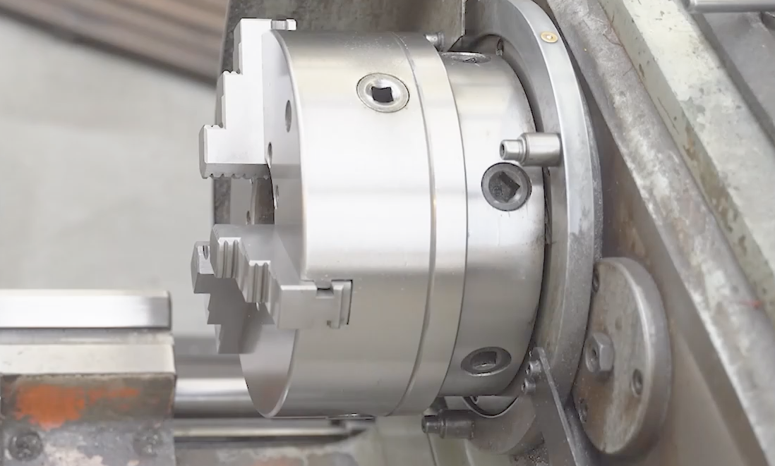 Na grafice przedstawiono wrzeciono tokarki w postaci precyzyjnie ułożyskowanego elementu w kształcie wału z otworem osiowym. Służy do zamocowania obrabianego przedmiotu – pośrodku wału znajduje się okrągły otwór oraz trzy metalowe uchwyty kształtem zbliżone do schodków ułożone równomiernie na obwodzie podstawy wału. Element ten przymocowany jest do korpusu maszyny. 