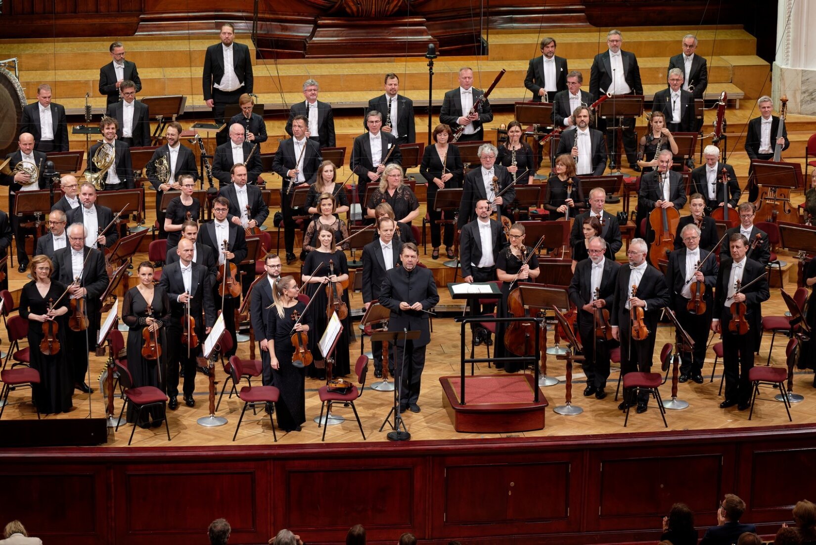Fotografia przedstawia grupę muzyków stojących na scenie sali koncertowej. Orkiestra składa się z mężczyzn i kobiet, mężczyźni ubrani są w czarne spodnie, czarne fraki i białe koszule, kobiety są w czarnych sukniach. Muzycy stoją, przodem do widowni, w rękach trzymają instrumenty, wśród nich stoi dyrygent z batutą w dłoniach. Przód sceny zajmuje pulpit dyrygenta, po obu bokach widoczne są krzesełka, na których siadają muzycy. Przed każdym muzykiem stoi pulpit z nutami.