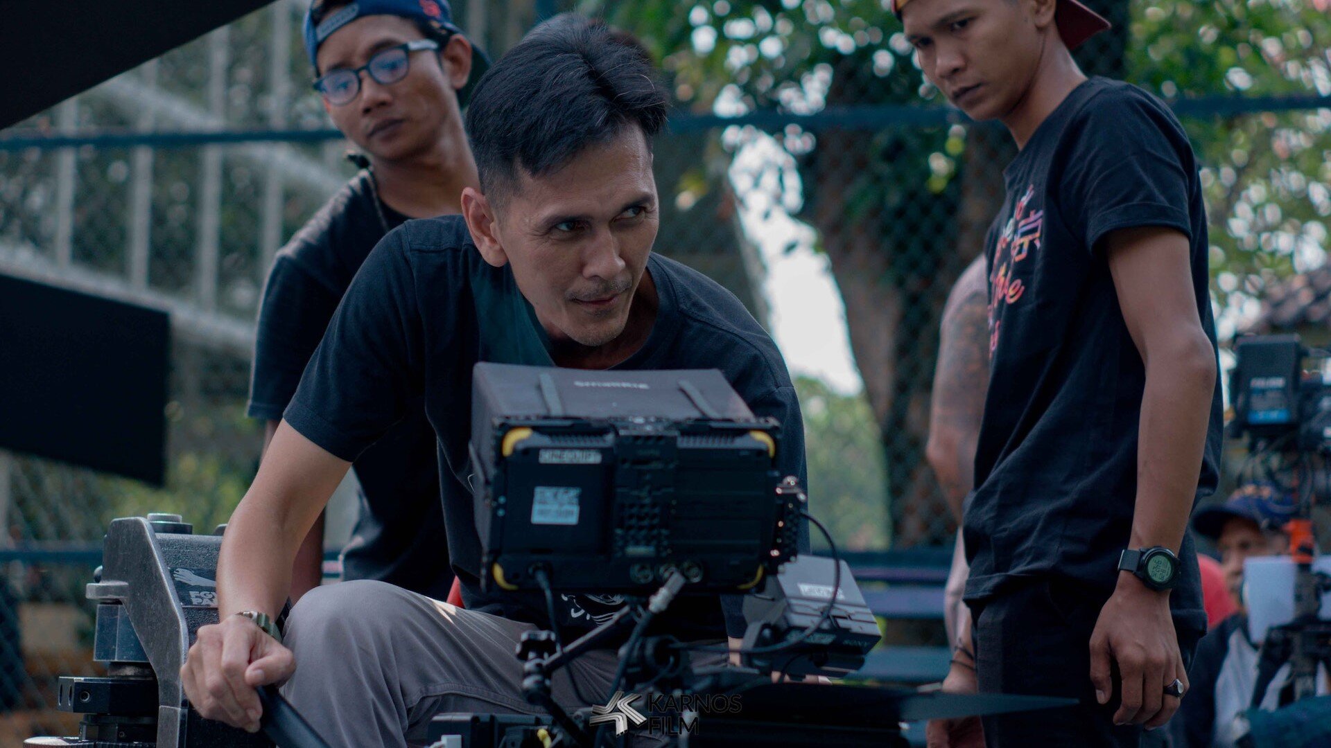 Zdjęcie przedstawia mężczyznę, który spogląda w ekran kamery na planie filmowym. Za nim stoi jeszcze dwóch mężczyzn.