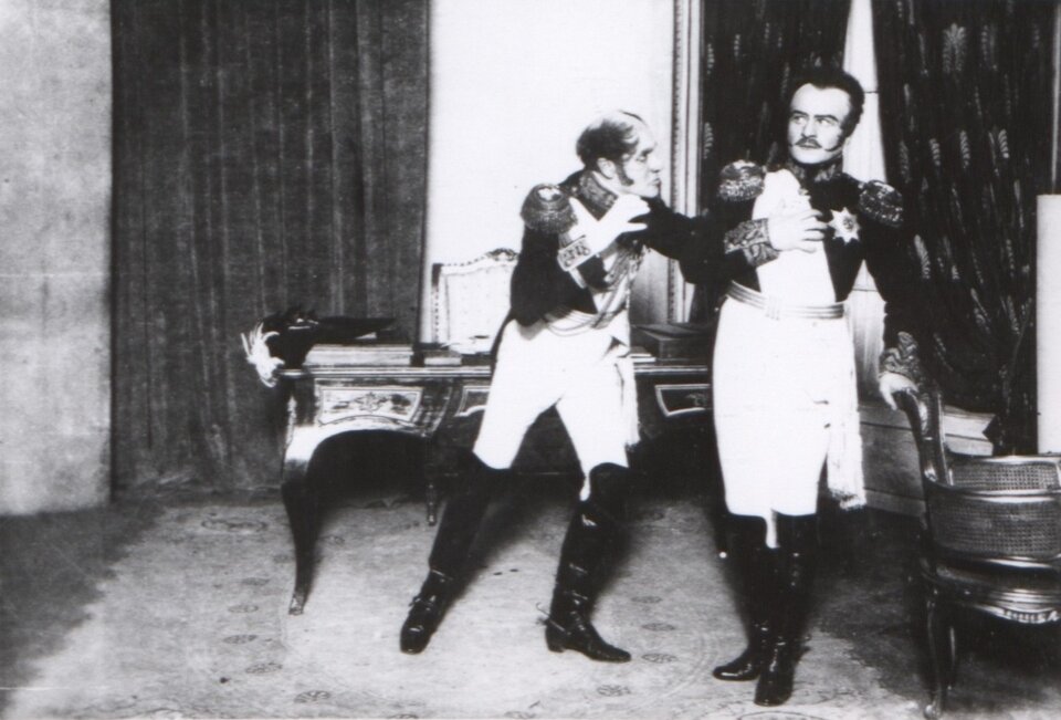 Ilustracja przedstawia scenę ze spektaklu „Kordian” w Teatrze Polskim w Warszawie. Po lewej stronie stoi wielki książę Konstanty. Ma on łysinę na głowie i baki. Jest ubrany w koszulę, mundur i spodnie. Na nogach ma długie buty za kolana. Patrzy w stronę cara. Gestykuluje obiema rękami. Za jego plecami znajduje się biurko, na którym po lewej stronie leży zamknięta księga, a na niej czapka. Za księgą leży notes. Za biurkiem, po lewej stronie znajduje się kotara. Przy biurku stoi krzesło. Car Mikołaj I stoi z prawej strony. Ma wąsy i włosy zaczesane do tyłu z bakami. Jest ubrany w koszulę, mundur z szarfą na lewym ramieniu. Ma na sobie spodnie i długie buty za kolana. Prawą dłonią dotyka stojącego krzesła. Lewą trzyma na sercu. Za plecami cara znajduje się okno z odsłoniętymi zasłonami. 