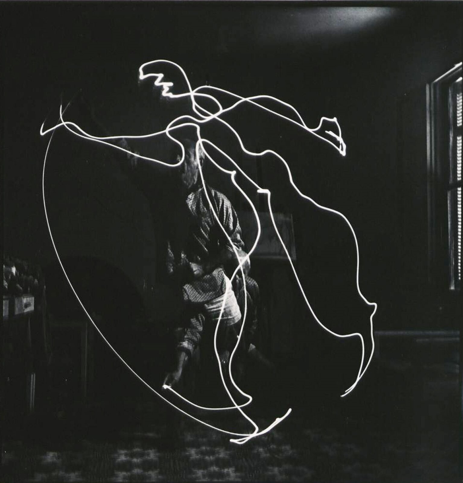 Ilustracja, czarno‑biała, przedstawia postać starszego mężczyzny z brodą. Mężczyzna wykonuje zamaszyste ruchy w powietrzu trzymając źródło światła. Dzięki długiemu czasowi naświetlania, na zdjęciu znajduje się długa smuga białego koloru. Jest to trasa ruchu światła wydobywającego się z soczewki latarki. Postać została zatrzymana w kadrze w dwóch pozach jednocześnie, stojąc z podniesioną ręką oraz przykucnięty z ręką wyciągniętą do przodu. Postacie nakładają się na siebie. Od dłoni biegną łukowate, świetliste linie.