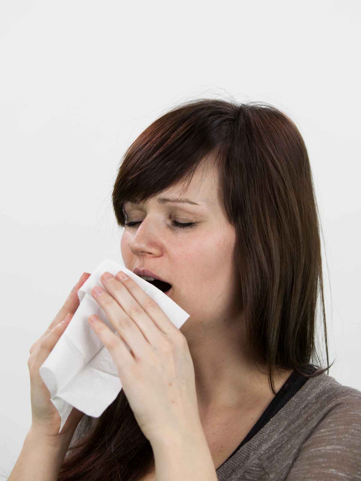 Zdjęcie przedstawia kichającą kobietę. Twarz zwrócona w lewo, oczy zamknięte, otwarte usta częściowo przysłonięte chusteczką trzymaną w rękach. Uchwycony moment na ułamek sekundy przed samym kichnięciem.