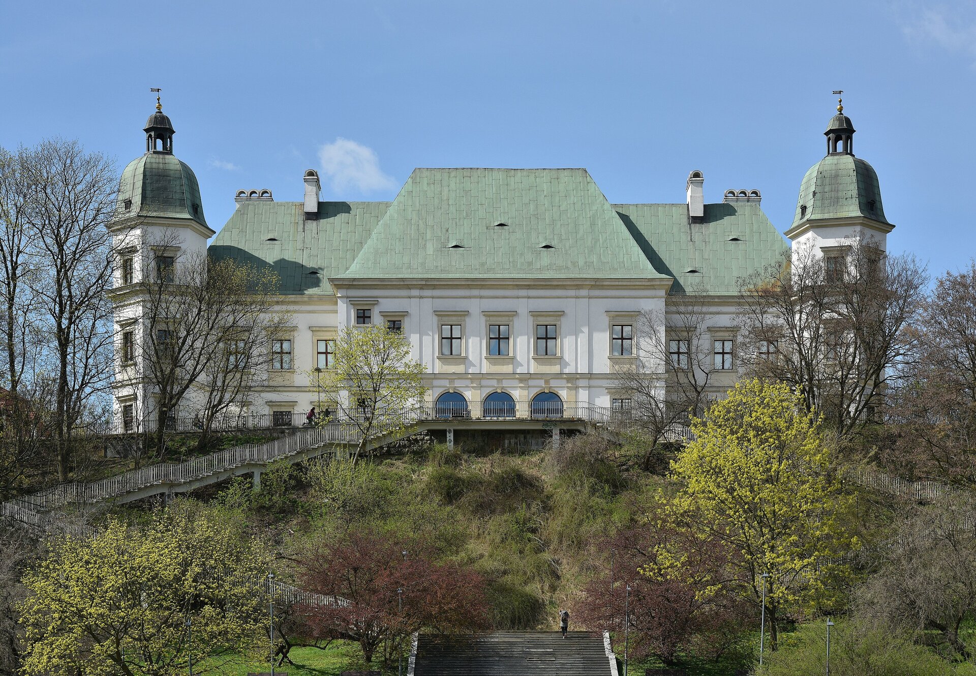 Zdjęcie przedstawia niewielki, biały budynek z zielonym dachem. Na pierwszym planie widoczny jest ogród otaczający zamek. Po bokach budynku znajdują się dwie wieże. Ich dach nie są strzeliste, tylko okrągłe.