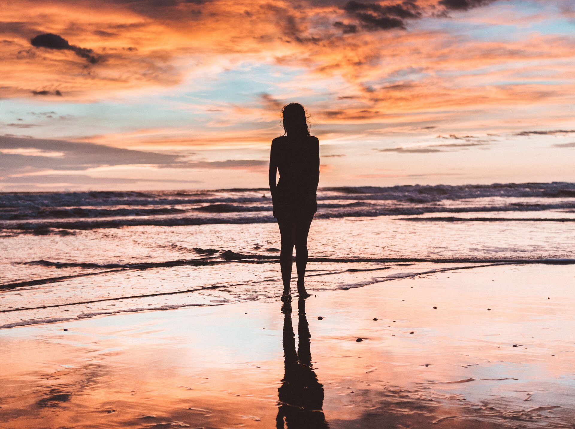 Rys. a. Zdjęcie przedstawia sylwetkę dziewczyny stojącej na piaszczystej plaży tuż przy brzegu morza. Niewielkie fale morskie obmywają jej bose stopy. 