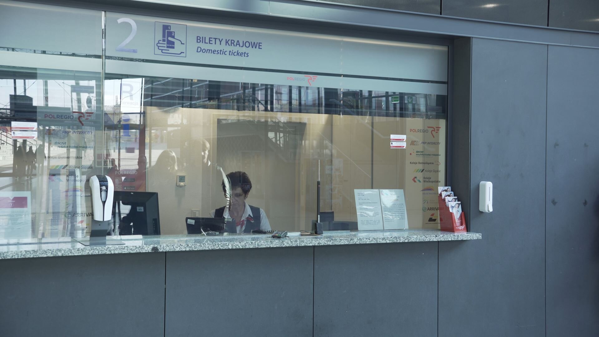 Zdjęcie przedstawia kasjera biletowego. Widoczne jest okienko kasy biletowej podpisane jako okienko numer dwa, bilety krajowe. Za szybą widać siedzącą przy komputerze kobietę w stroju kasjera biletowego.