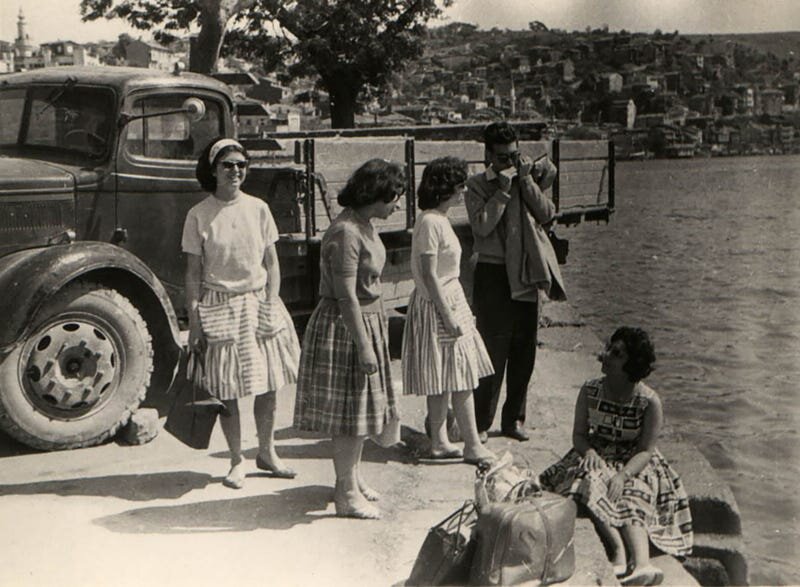 Zdjęcie przedstawia grupę ludzi. Głównie to kobiety w towarzystwie jednego mężczyzny. Trzyma cos blisko ust, może przygrywa na harmonijce. Dziewczyny ubrane w sukienki do kolan z krótkim rękawem, na nosie okulary przeciwsłoneczne. Pogoda dopisuje. W stóp trzymają walizki. Znajdują się przy rzece lub zatoce. Za ich plecami stoi auto z naczepą. Na ostatnim planie zabudowa miejska oraz meczet. 