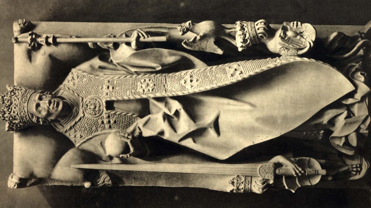 Ilustracja przedstawia nagrobek Kazimierza Jagiellończyka widziany od góry. To rzeźba postaci jednocześnie leżącej i stojącej. Ramiona i głowa spoczywają na poduszce,  ale nogi opierają się na konsoli - takie ujęcie sugeruje, że postać stoi. Król jest ubrany w strój koronacyjny, na głowie ma koronę, w prawej dłoni jabłko, w lewej berło, na palcach pierścienie, u boku skierowany ostrzem do góry, wielki miecz. Na nagrobku znajdują się trzy herby: podwójny krzyż Jagiellonów, herb Polski i herb Habsburgów. Lew po lewej stronie trzyma równocześnie miecz króla. Nad głową lwa po stronie przeciwnej umieszczony jest orzeł w koronie, klejnot herbu Polski.