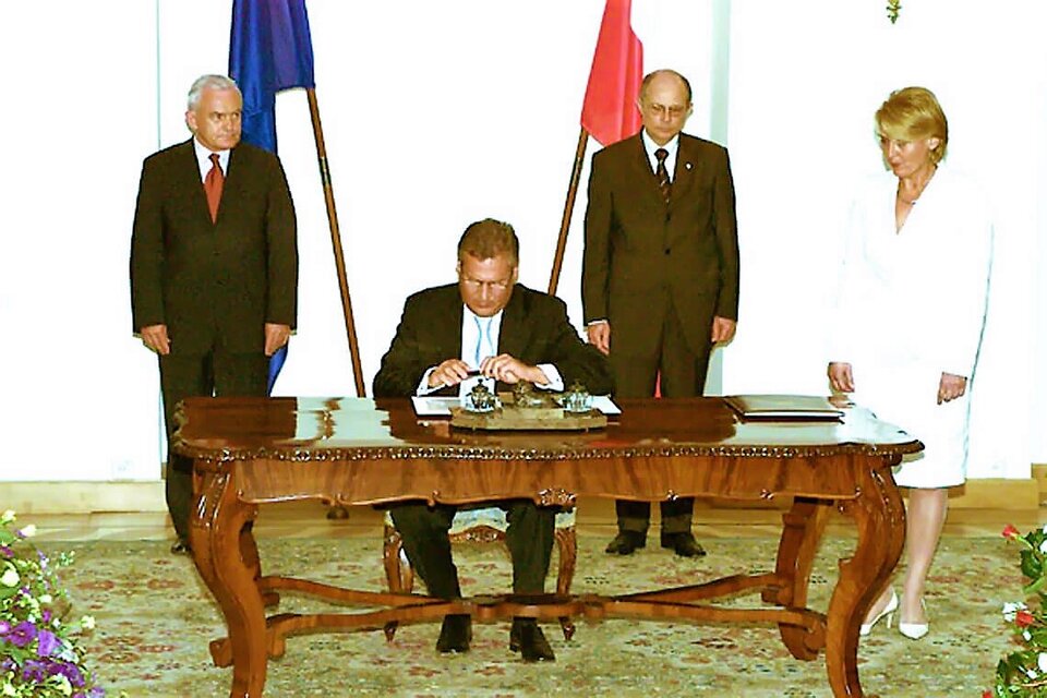 Zdjęcie przedstawia Aleksandra Kwaśniewskiego siedzącego przy stole i podpisującego dokumenty. U jego lewego boku stoi kobieta, a za plecami Leszek Miller i Marek Borowski. Obok nich stoją flagi Polski i Unii.