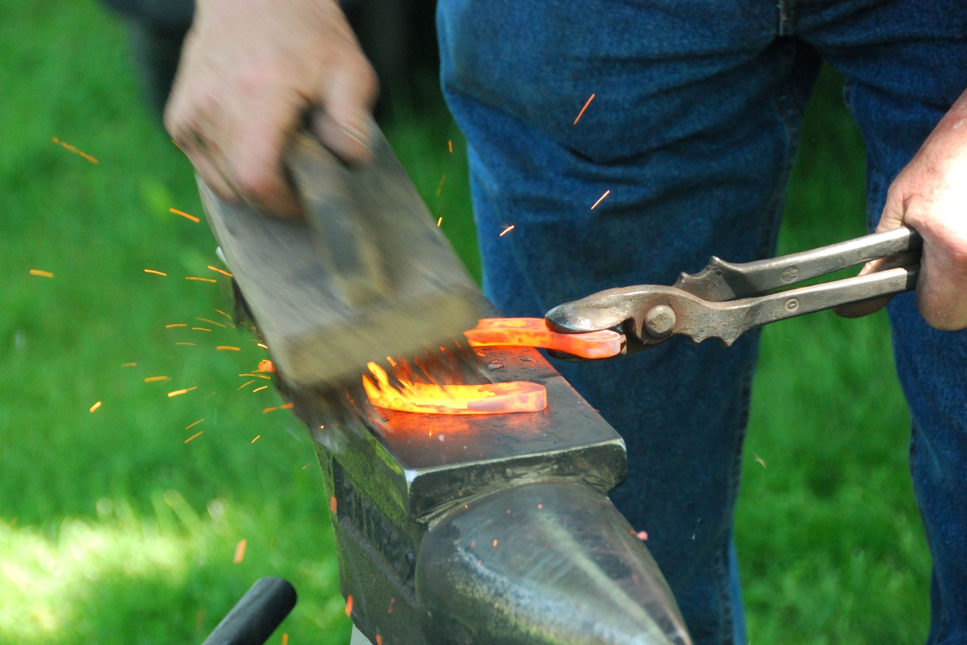 Fotografia przedstawia moment wykuwania podkowy. Na fotografii widoczne są ręce kowala. W lewej trzymane szczypce przytrzymujące rozżarzoną do czerwoności podkowę, opartą o kowadło. W prawej trzymany młot kowalski uderzający o podkowę. Widoczny rozprysk iskier powstały na skutek uderzenia. Na dalszym planie widoczne nogi kowala. W tle zielony trawnik.