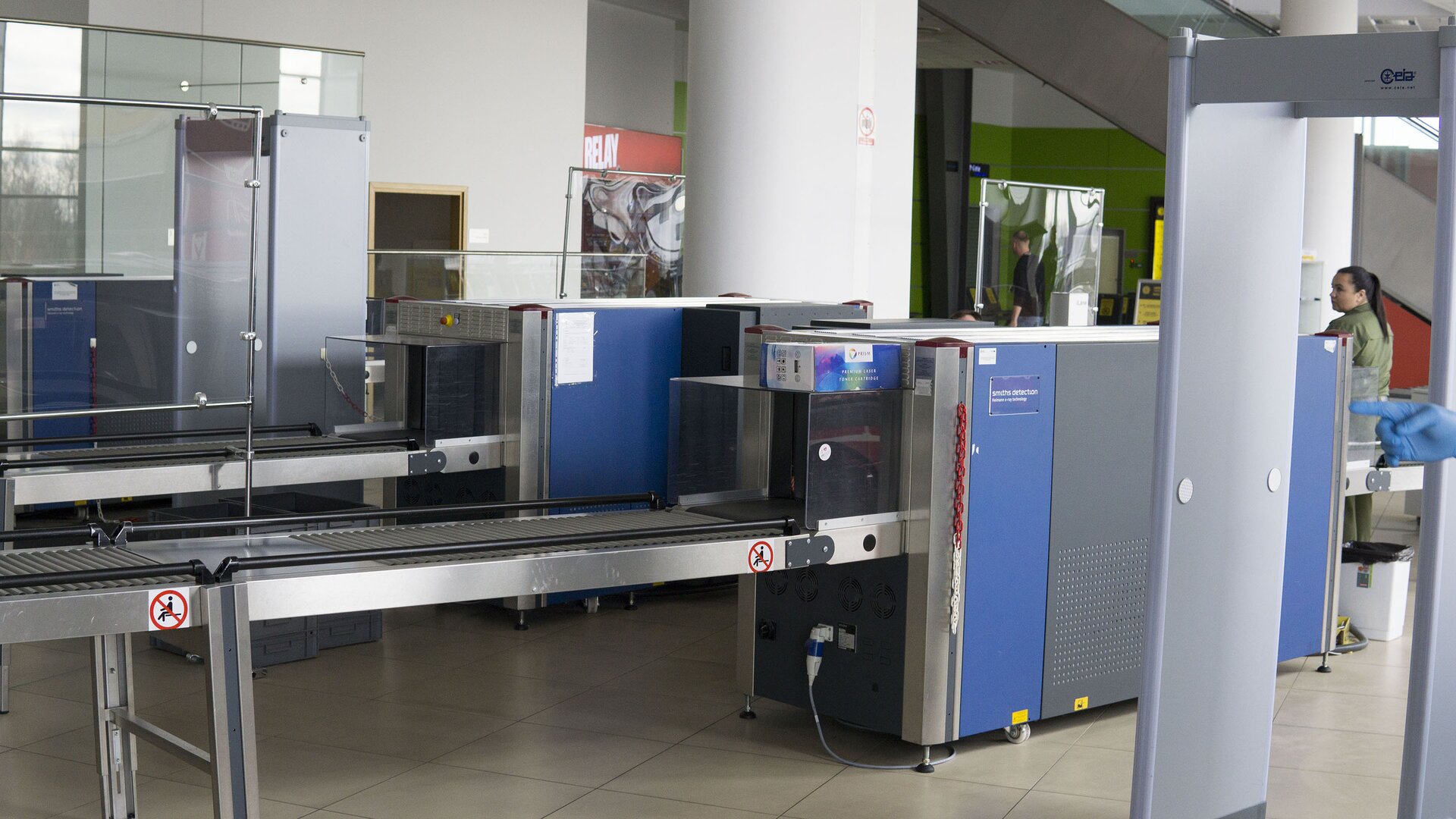 Zdjęcie przedstawia urządzenie do wykrywania materiałów wybuchowych, które stoi w punkcie kontroli lotniska. Urządzenia składa się zasadniczo z dwóch głównych części: prześwietlającej oraz taśmy transportującej bagaż. Część prześwietlająca bagaż to prostopadłościan o wysokości około półtora metra i długości około dwóch metrów. W przedniej i tylnej ścianie wycięte są prostokątne otwory, przez które przechodzi taśma. Na niej bagaże wjeżdżają do części w kształcie prostopadłościanu, gdzie są prześwietlane. Taśma ustawiona jest na nogach i ma kilka metrów długości. Mniej więcej na środku jej długości stoi prostopadłościenna część prześwietlająca. Na bocznej ramie taśmy umieszczono naklejki z ostrzeżeniem, że nie wolno siadać na taśmie.