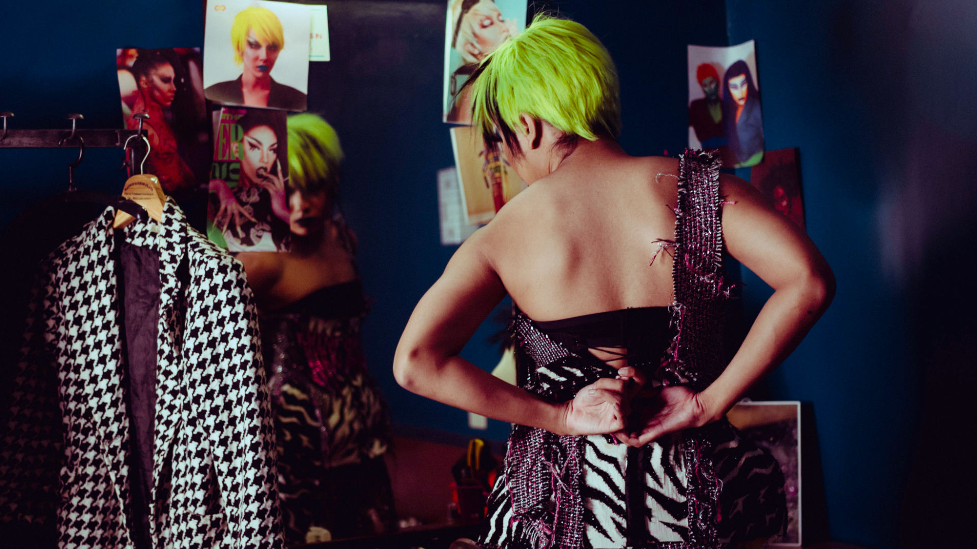 Zdjęcie przedstawia kobietę, która przymierza  kostium i przegląda się w lustrze. Obok po lewej na wieszaku wisi inny kostium.