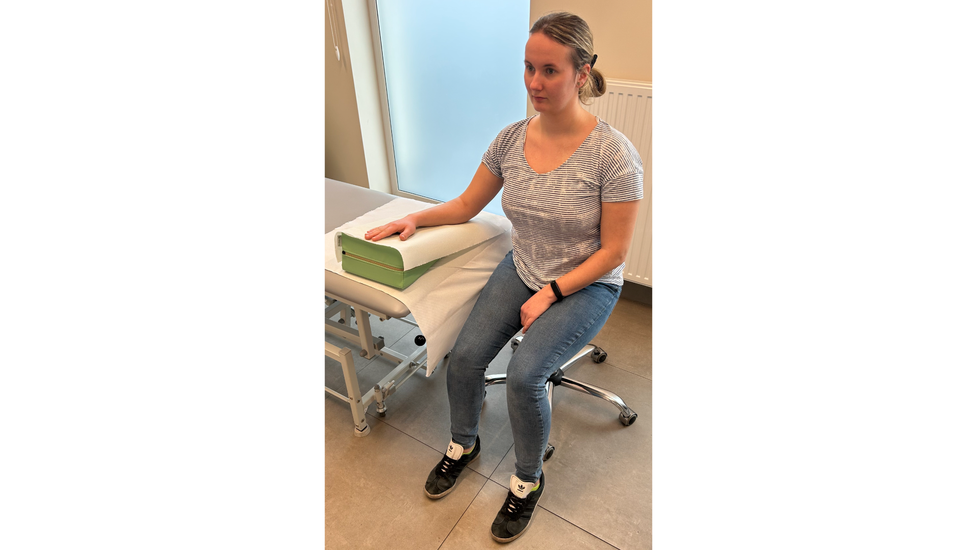 Zdjęcie przedstawia pacjenta siedzącego na krześle obok stołu do masażu. Prawa ręka, oparta na klinie trójkątnym leżącym na stole do masażu. Klin oraz stół znajdują się na wysokości klatki piersiowej.