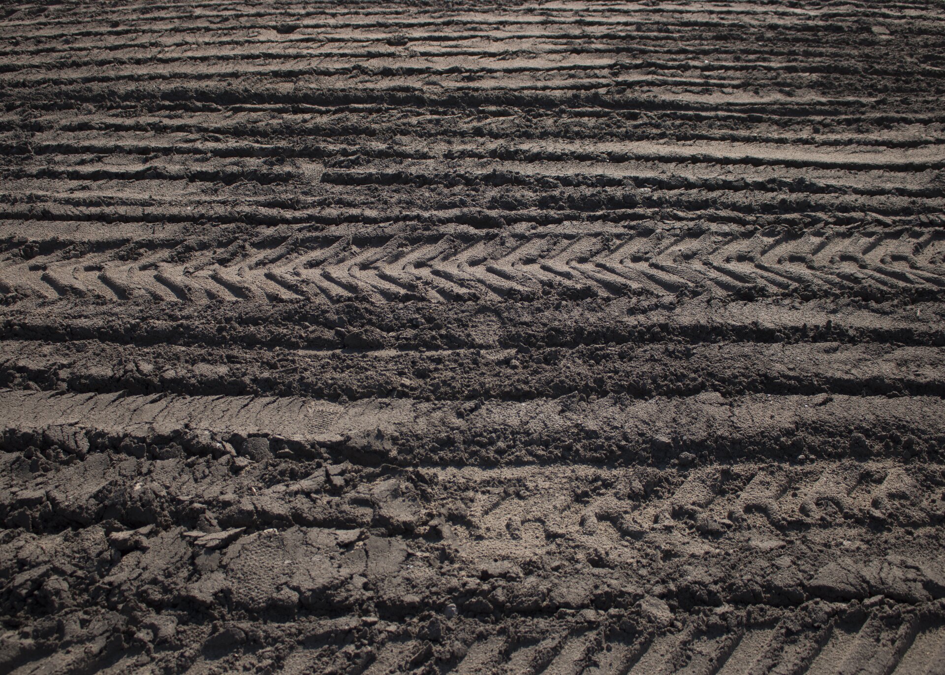 Ilustracja przedstawia ślady pojazdu na ciemnobrązowej ziemi. Na fotografii widoczne są odbite na podłożu odciski bieżnika opon samochodowych, ułożone w poziome pasy o różnych szerokościach.