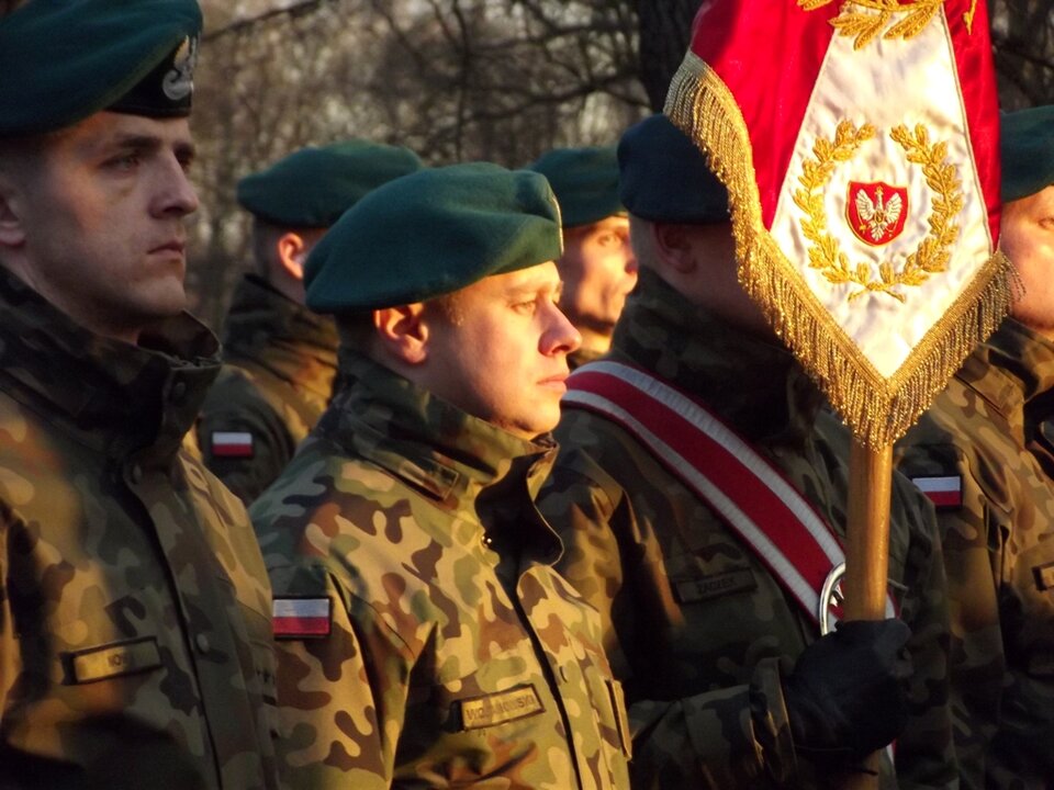 Kliknij, aby powiększyć Fotografia żołnierzy polskich. Ubrani są w mundury moro, na głowie mają berety wojskowe. Jeden z nich trzyma przed sobą sztandar.