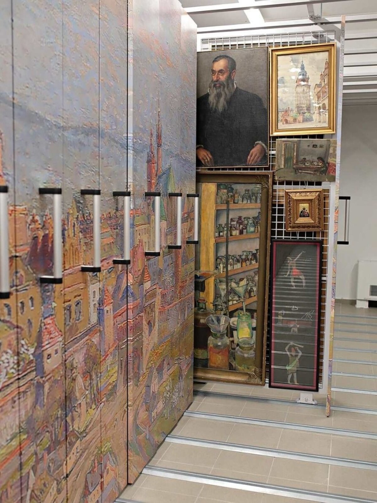 Na zdjęciu zbiór obrazów. Po lewej stronie fotografii znajduje się szafa, na której drzwiach znajduje się obraz miasta. Szafa składa się z ośmiu, wysuwanych drzwi. Na środku fotografii przedstawiono sześć obrazów, które wiszą w dwóch kolumnach na metalowych drzwiach szafy. Pierwsza "kolumna" to dwa obrazy: Obraz mężczyzny w podeszłym wieku o krótkich włosach oraz długiej i bujnej siwej brodzie, który jest ubrany w czarny płaszcz. Drugi obraz umieszczony został pod omawianym portretem i przedstawiaj półkę z różnymi słoiczkami oraz butelkami, które są różnej wielkości. Na samym dole obrazu znajdują się trzy duże słoiki, w których umieszczono lejki. Druga kolumna dzieł zaczyna się od obrazu w złotej ramie. Przedstawiono na nim miejski rynek, gdzie znajduje się kamieniczka oraz wysoki budynek ze strzelistą wieżą. Poniżej wiszą kolejne obrazy, to jest: obraz przedstawiający pomieszczenie w domu, obraz Matki Boskiej z dzieciątkiem oraz podłużny obraz z dwiema postaciami. 