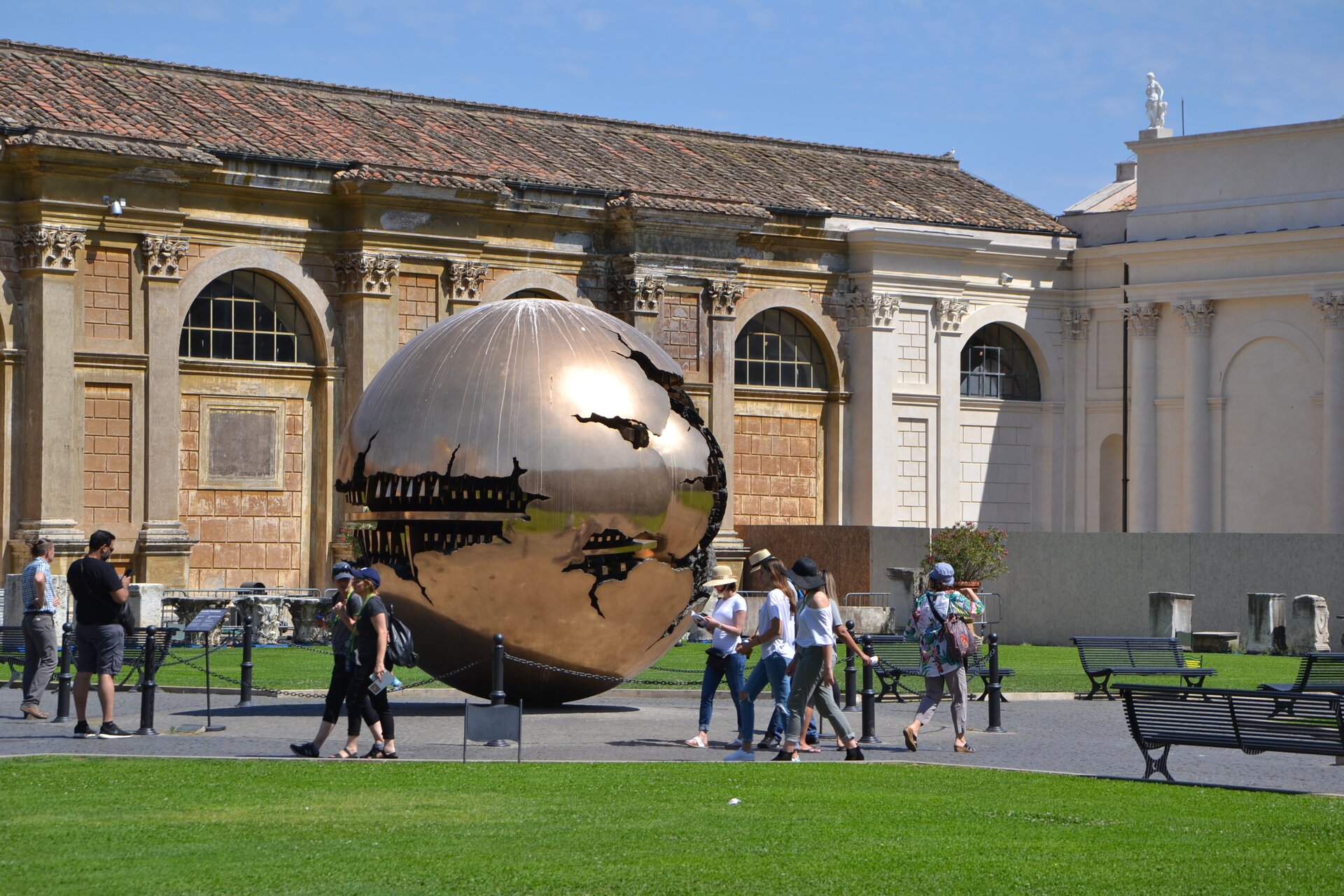 Ilustracja przedstawia fotografię fragmentu dziedzińca w Watykanie. Na pierwszym planie jest trawnik, wokoło którego są zabudowania. Pomiędzy nimi stoi ogromny pomnik w postaci kuli z wieloma nieregularnymi otworami. Wzdłuż trawnika spacerują ludzie.